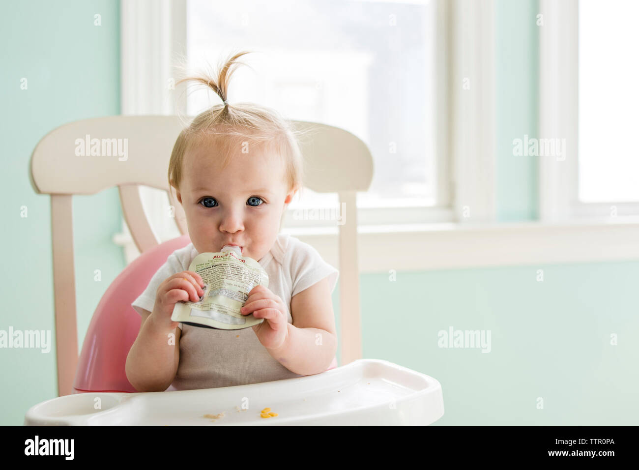 Portrait of cute baby girl eating apple sauce from pack while sitting on high chair at home Stock Photo