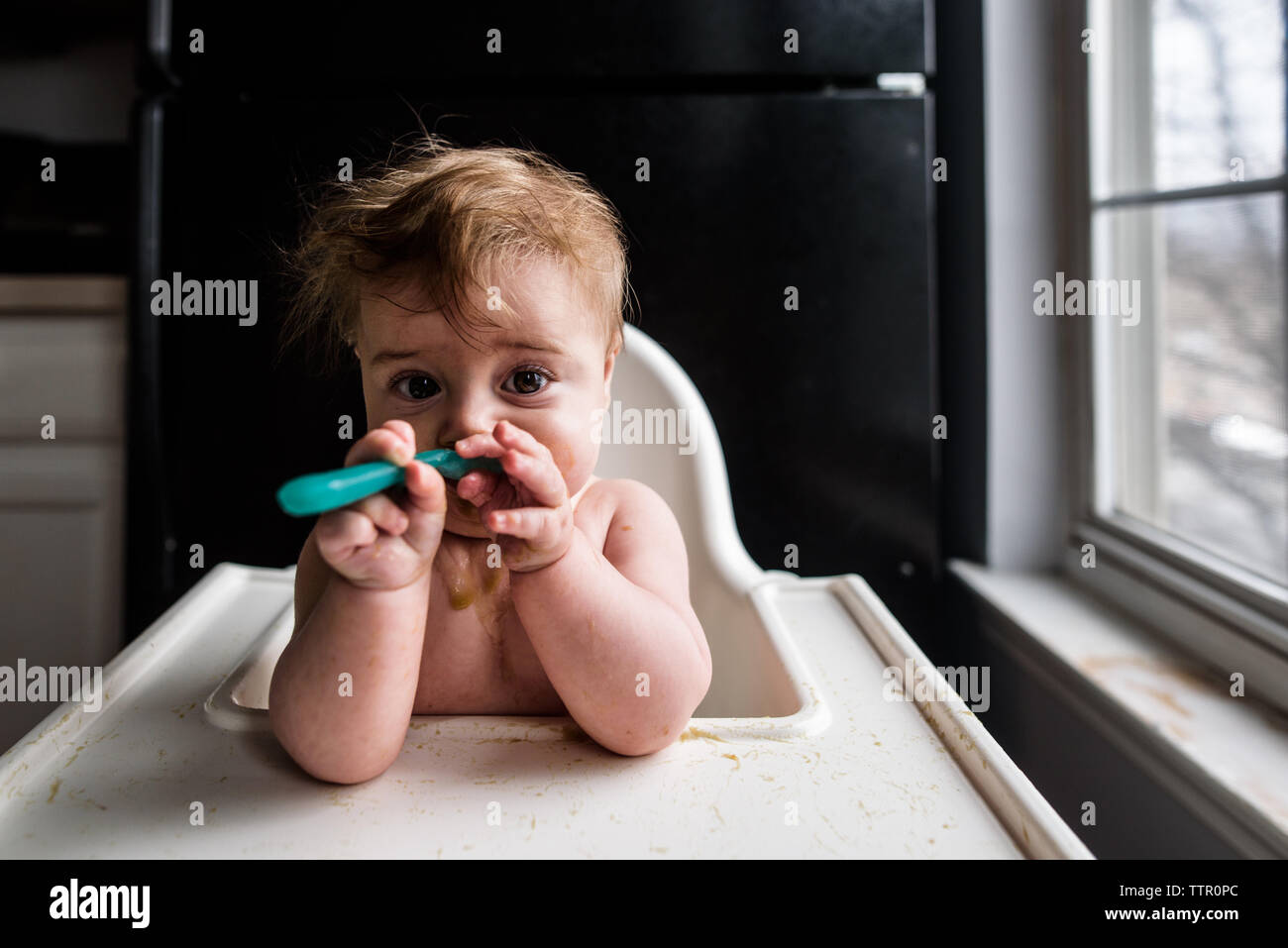 Close up shot of messy baby eating with a spoon in highchair Stock Photo