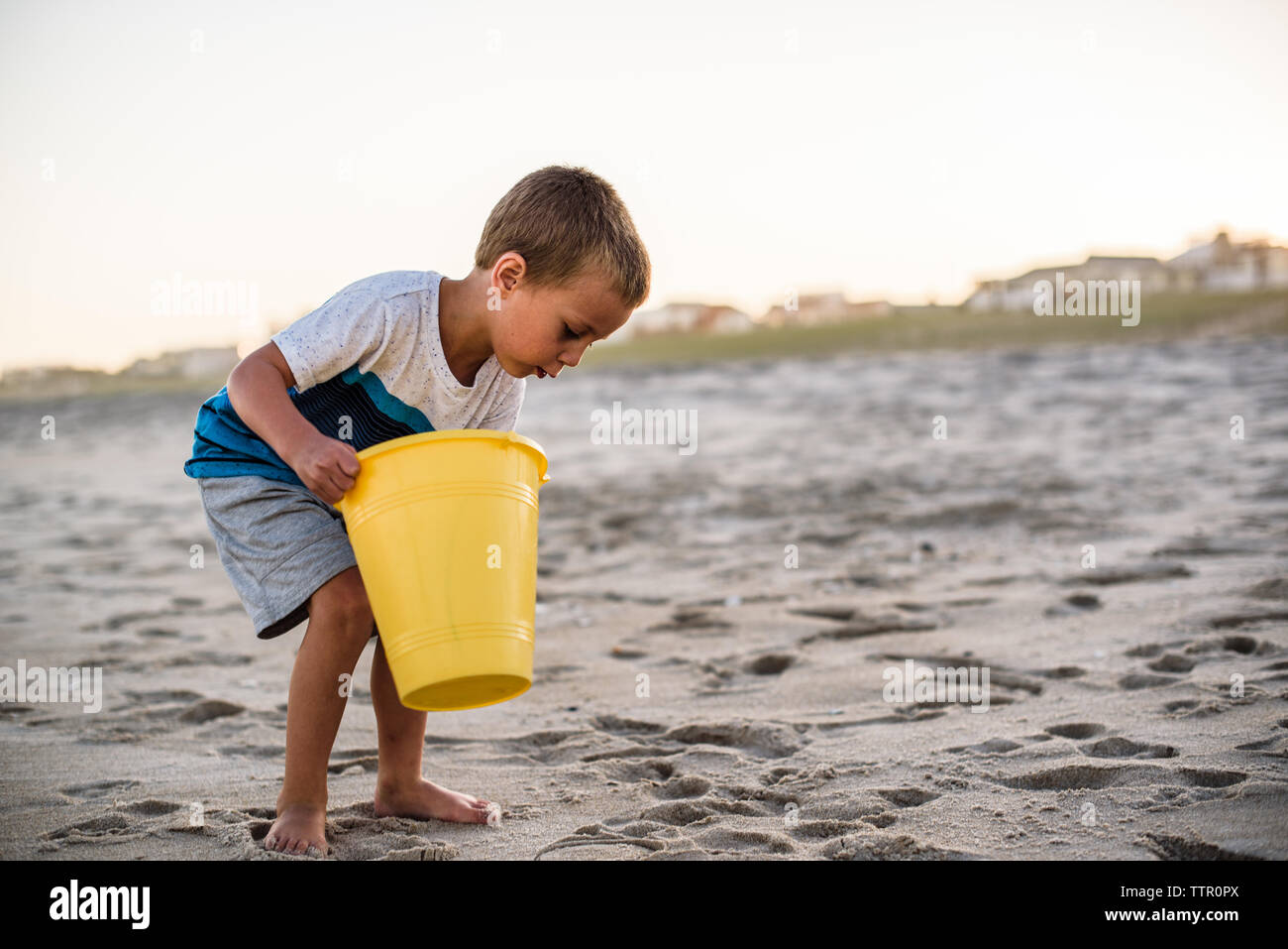 Boy on beach holding yellow bucket while searching the sand at sunset Stock Photo