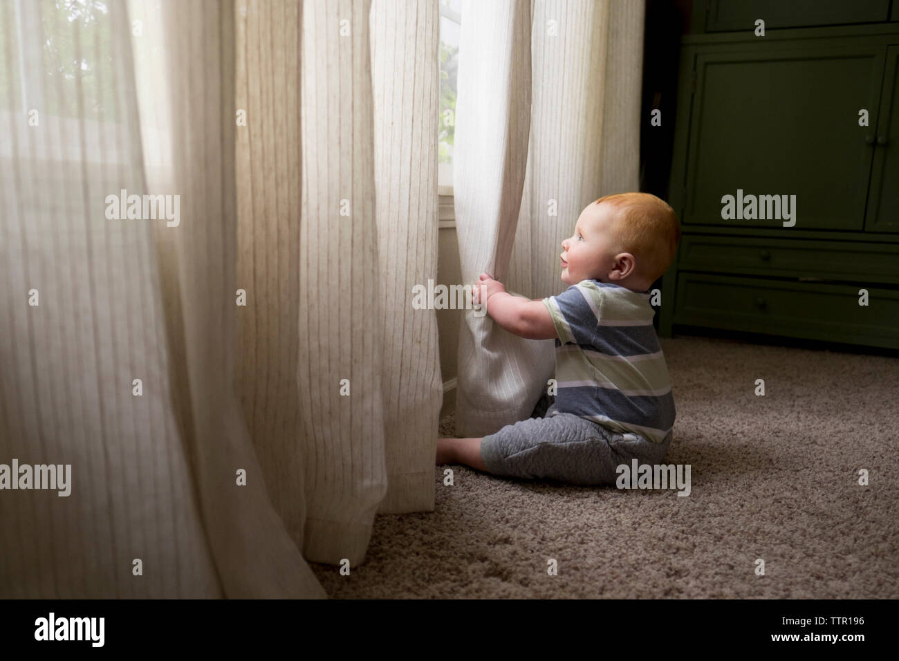 Side view of baby boy playing with curtains while sitting on carpet at ...