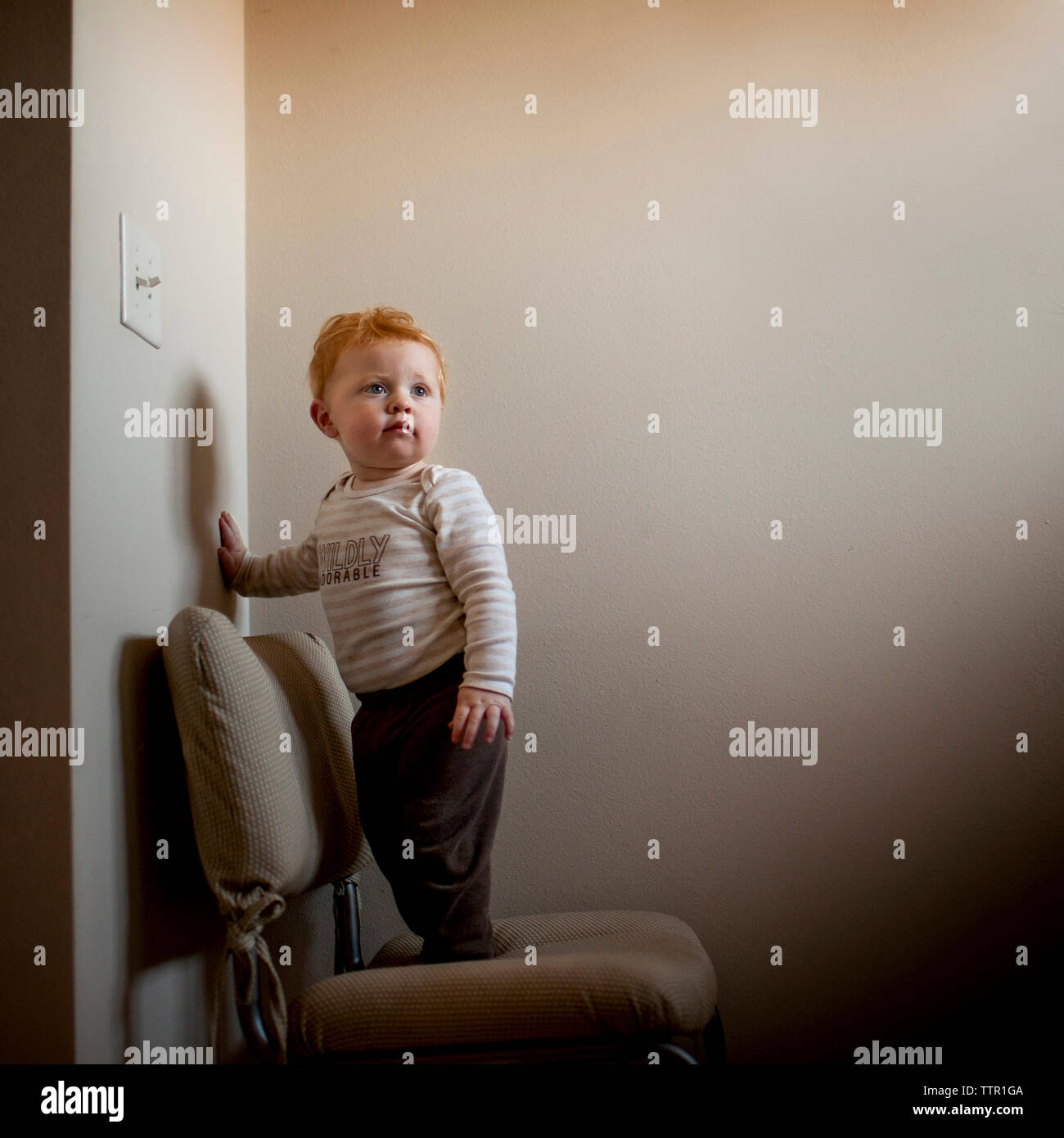 Cute baby boy looking away while standing on chair against wall at home Stock Photo
