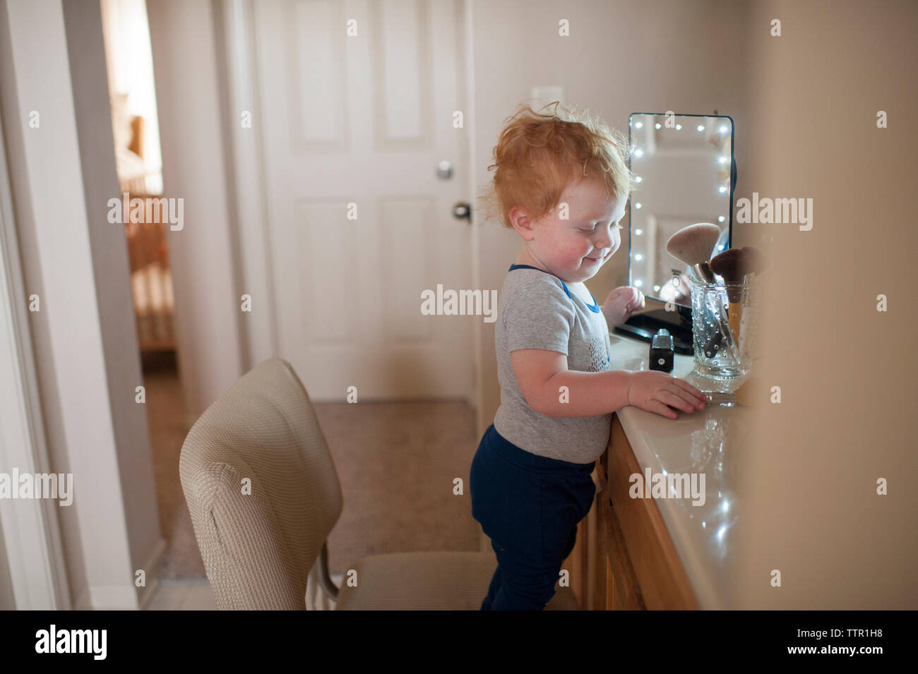 Side view of cute baby boy standing by beauty products on chair at home Stock Photo
