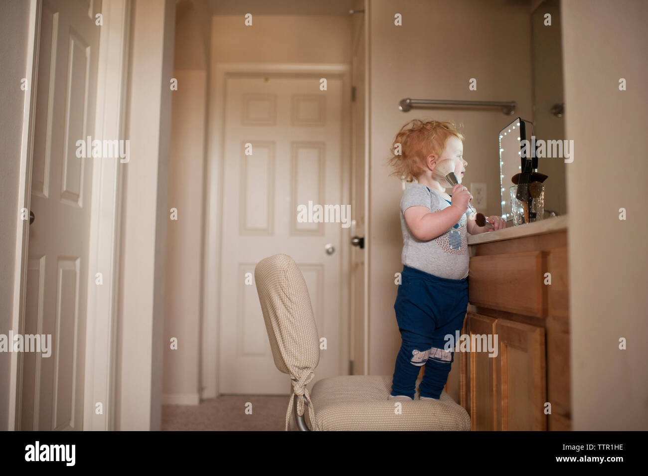 Cute baby boy applying make-up while standing on chair at home Stock Photo