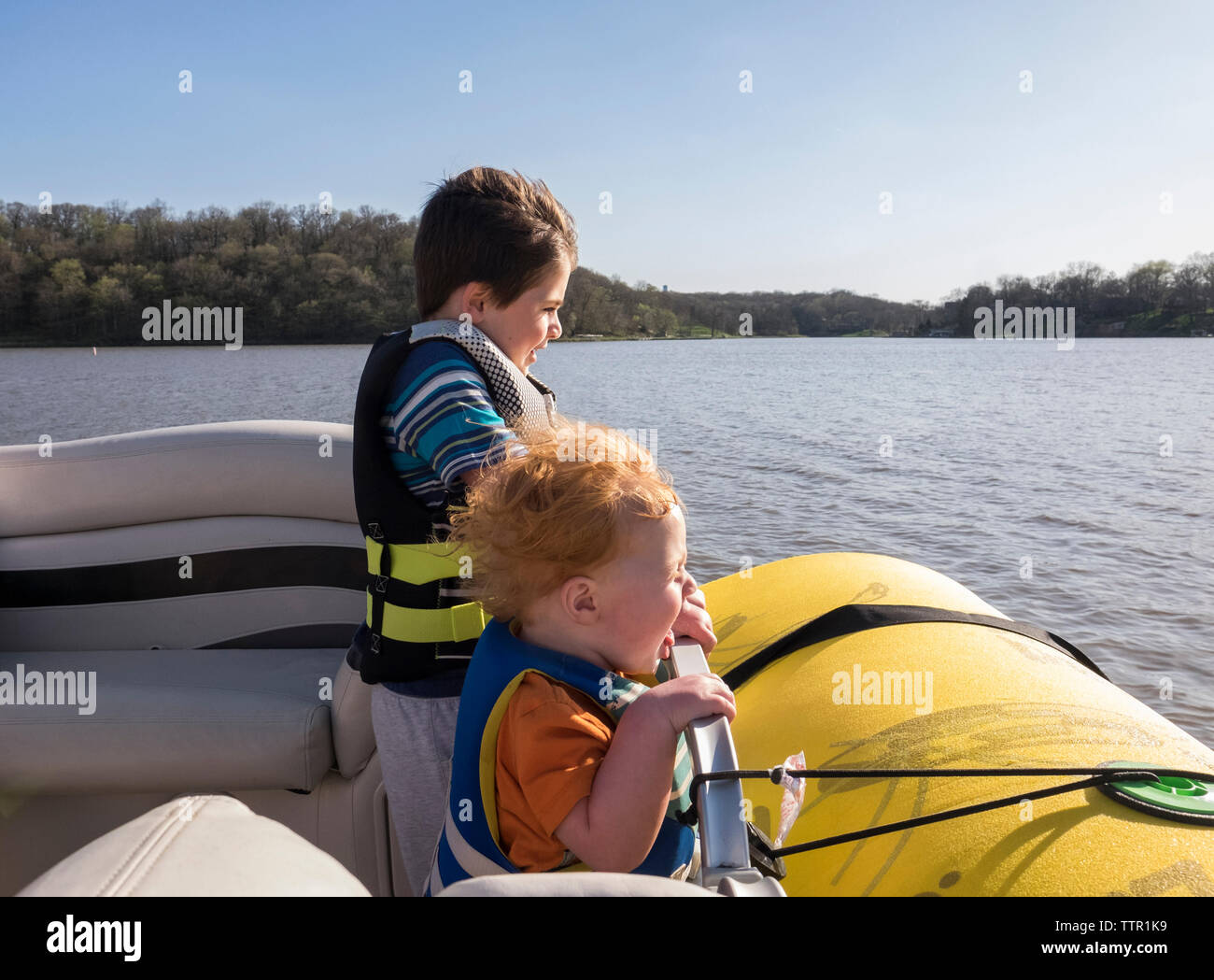 Happy brothers wearing life jackets while standing in boat on lake against clear sky during sunny day Stock Photo
