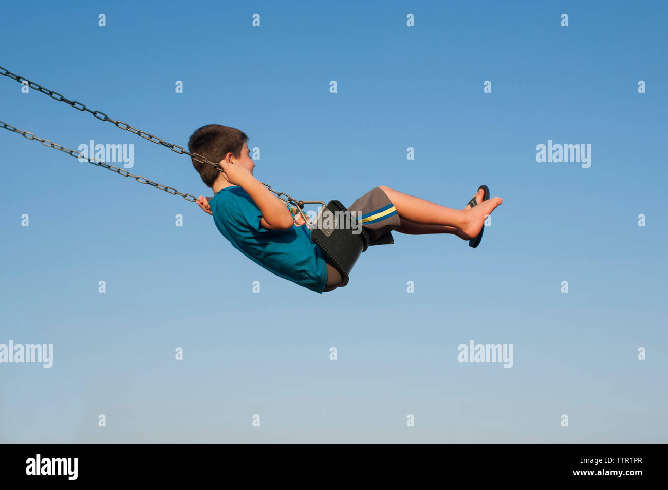 Side view of carefree boy swinging on swing against clear blue sky at playground Stock Photo
