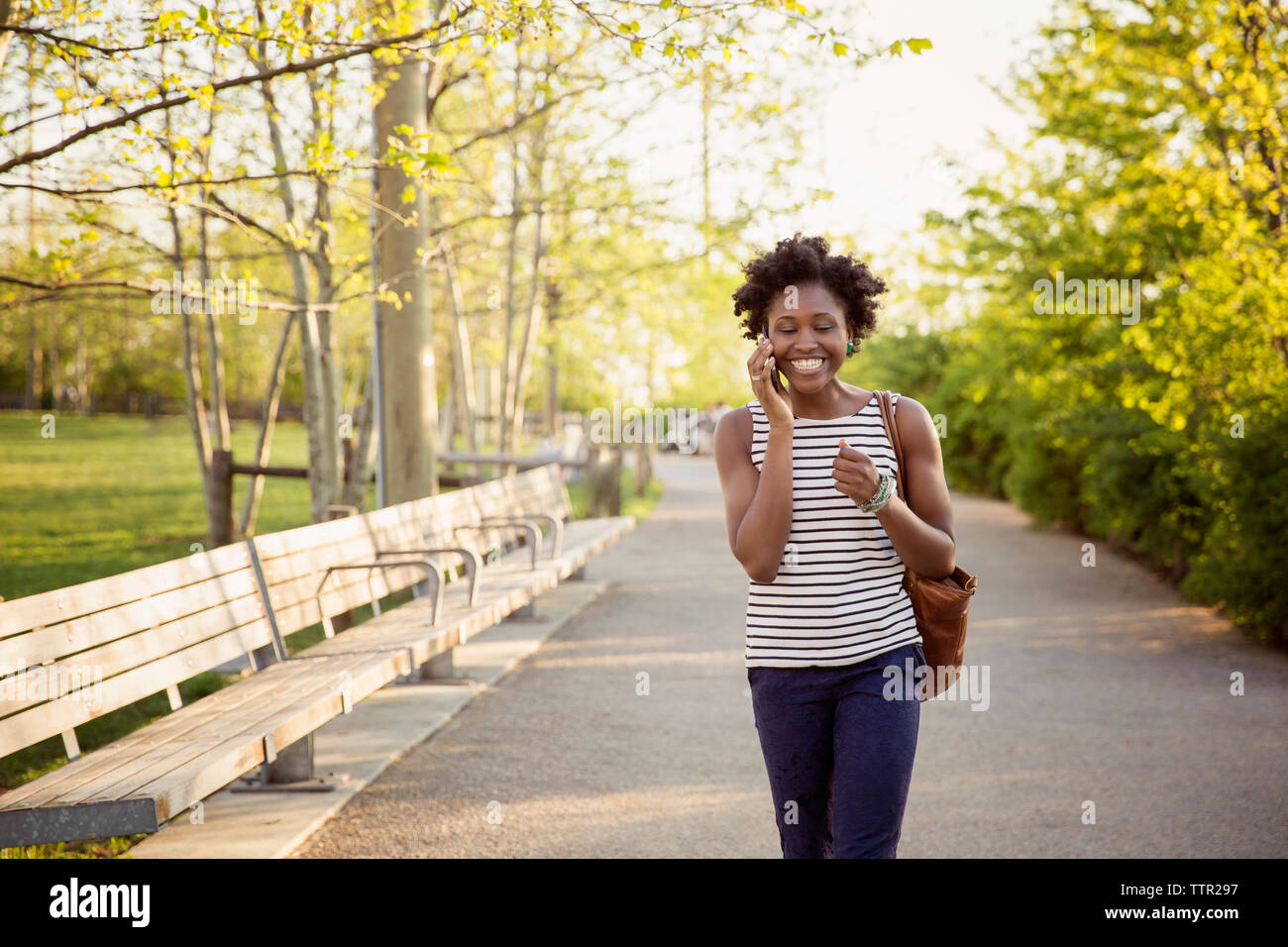 Cheerful woman using smart phone while walking on footpath in park Stock Photo