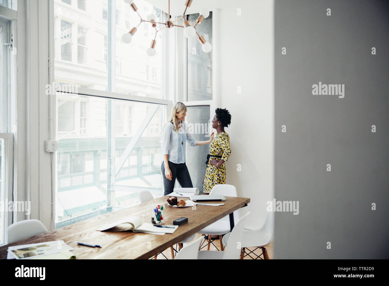 Side view of businesswomen talking while standing by window in board room Stock Photo