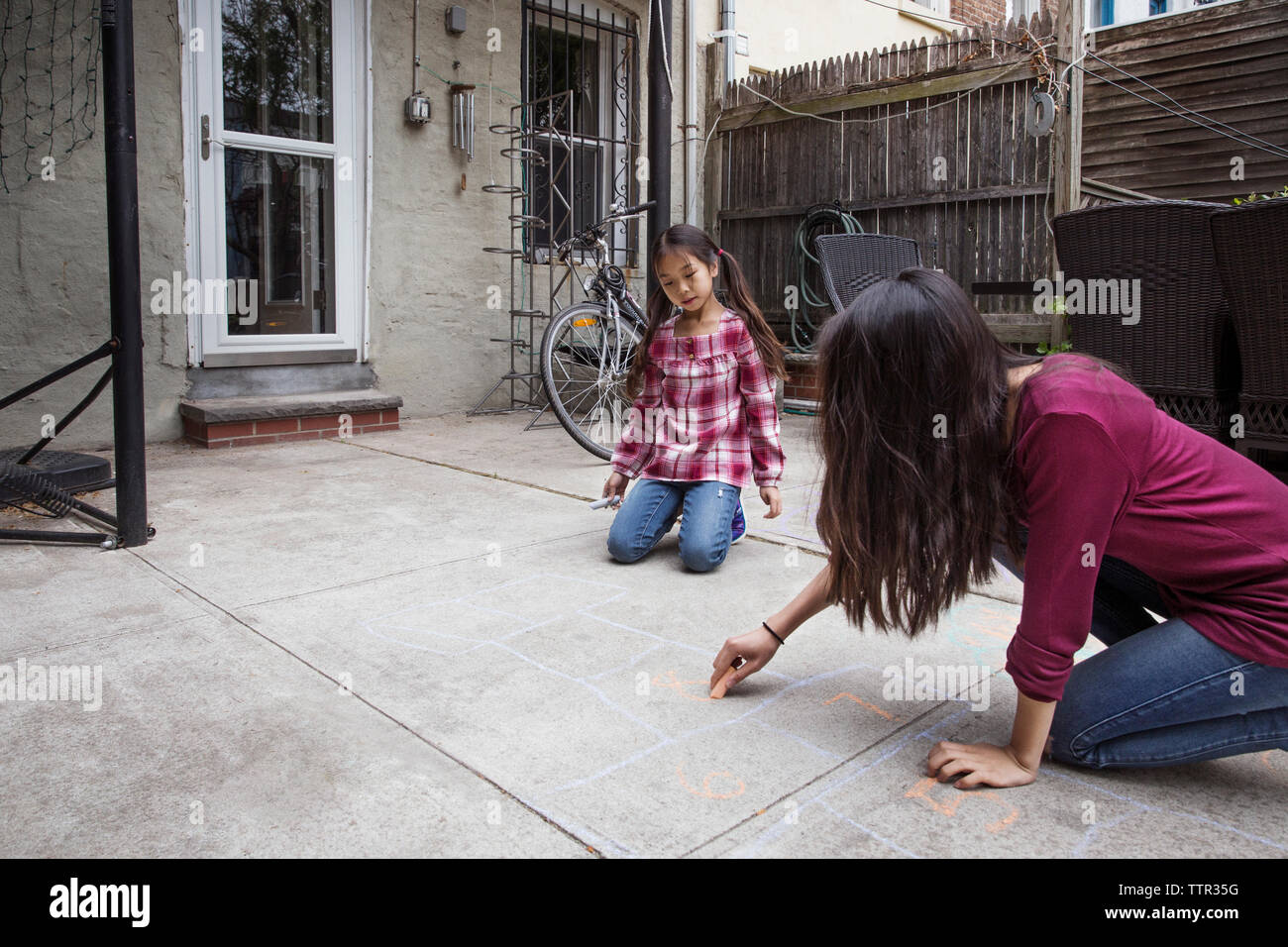 Girl looking at sister drawing hopscotch outside house Stock Photo