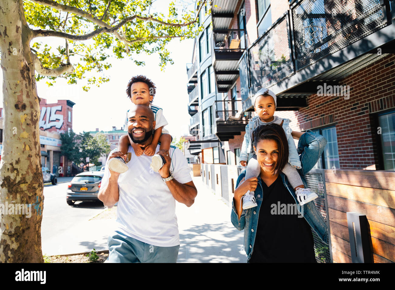 Portrait of happy parents carrying children on shoulders while walking on sidewalk in city Stock Photo