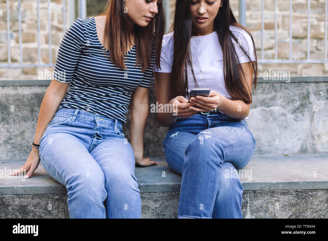 Female friends using mobile phone while sitting on steps against wall Stock Photo