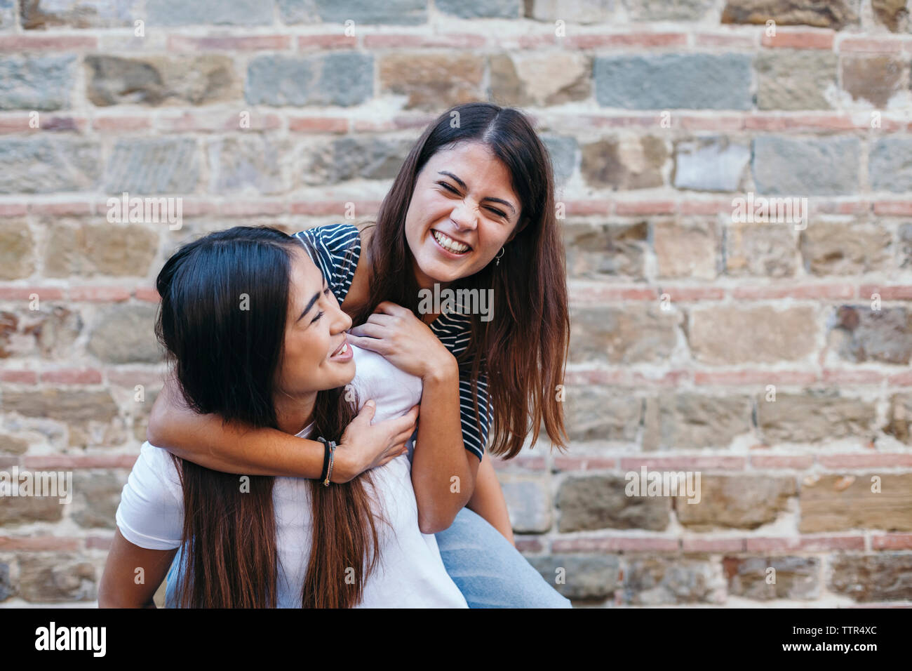 Woman piggybacking happy friend while standing by wall Stock Photo