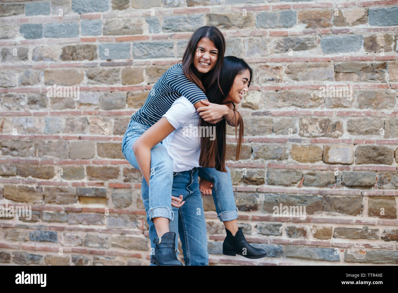 Happy woman piggybacking friend while standing by wall Stock Photo
