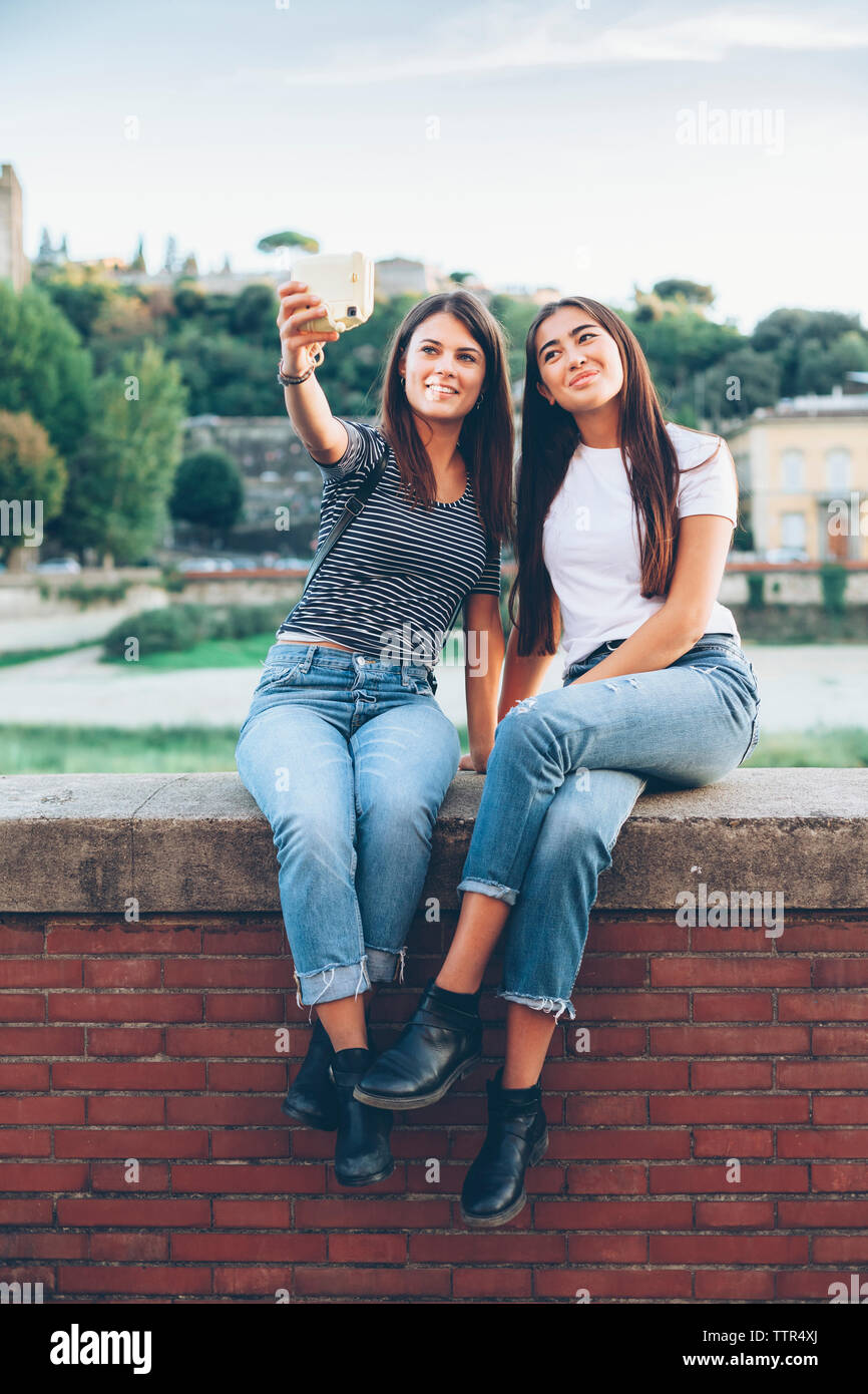 Full length of happy female friends taking selfie with instant camera while sitting on retaining wall at park Stock Photo