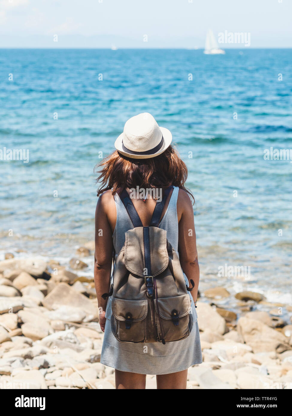 Rear view of woman with backpack looking at view while standing on rocky beach against sky during sunny day Stock Photo
