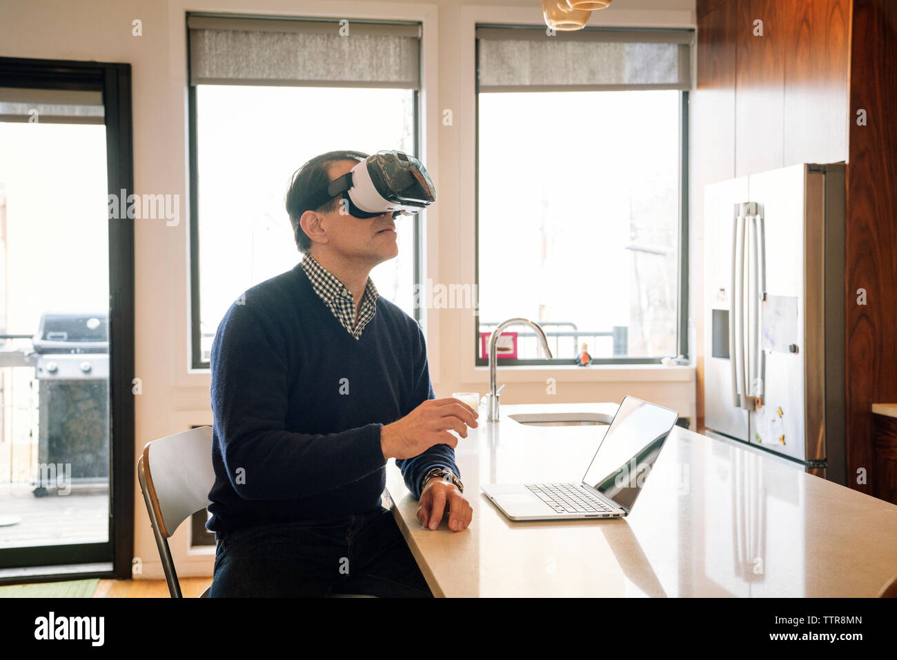 Man wearing virtual reality simulator sitting at table in kitchen Stock Photo