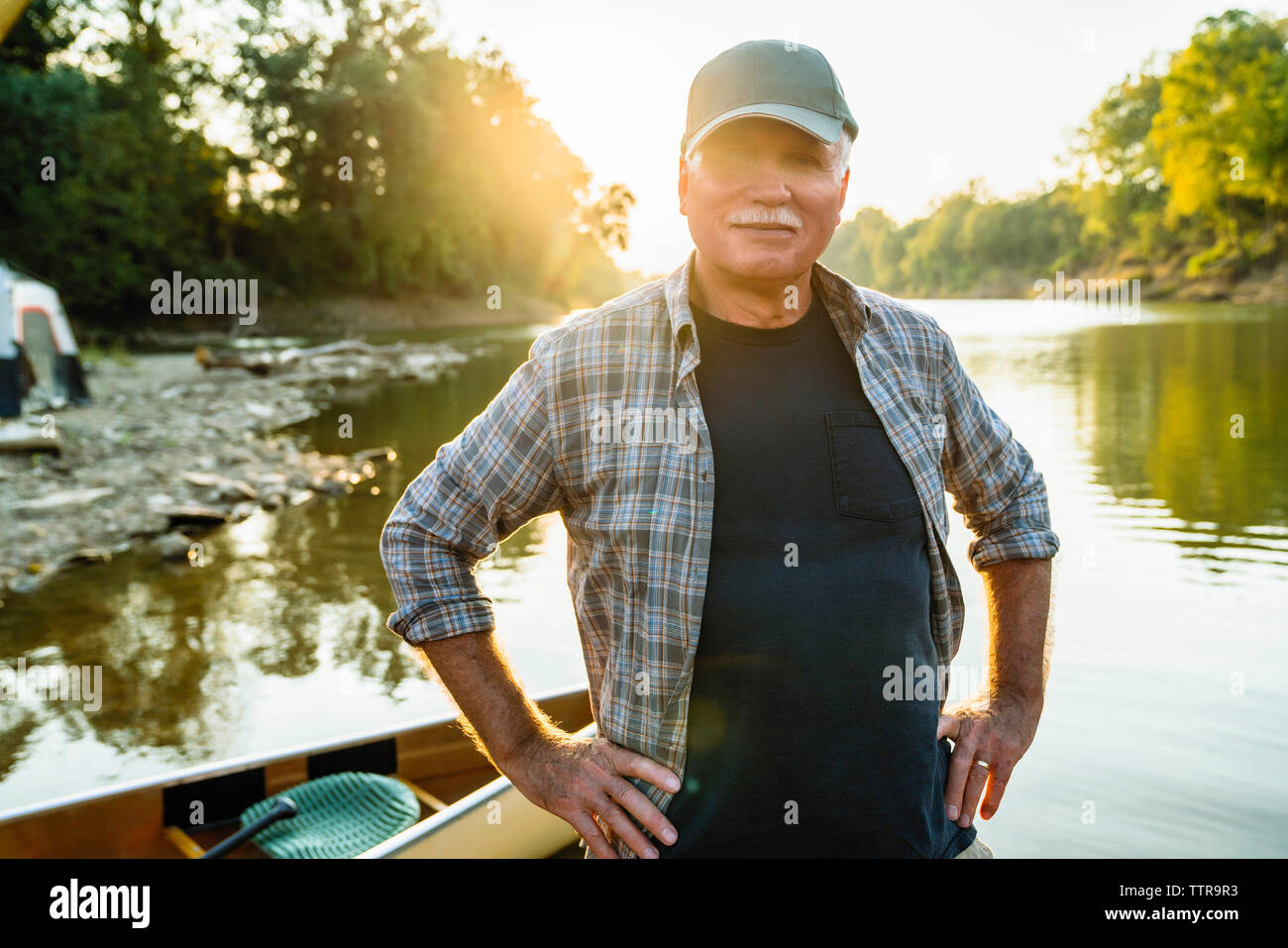 Portrait of senior man with hands on hip standing by boat at lakeshore during sunset Stock Photo