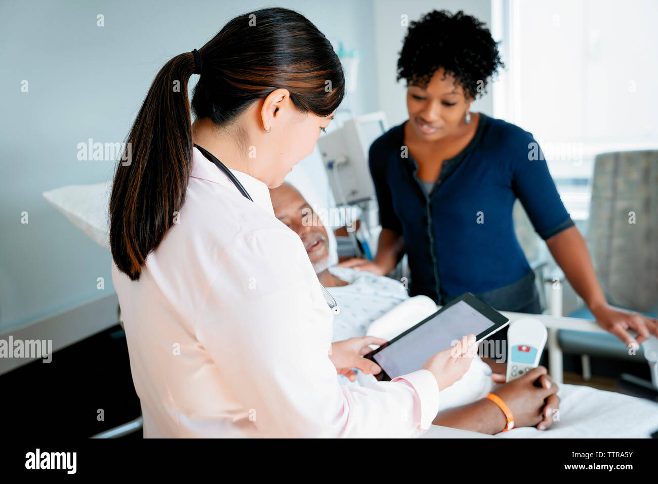 Female doctor looking at tablet computer while discussing with patient and woman in hospital ward Stock Photo