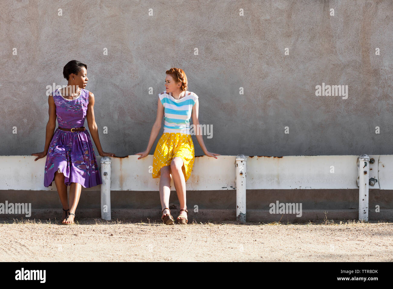 Multi-ethnic female friends talking while sitting on railing against wall Stock Photo
