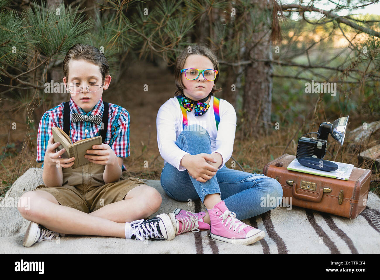 Siblings in old-fashioned costume sitting on blanket with antique camera at forest Stock Photo