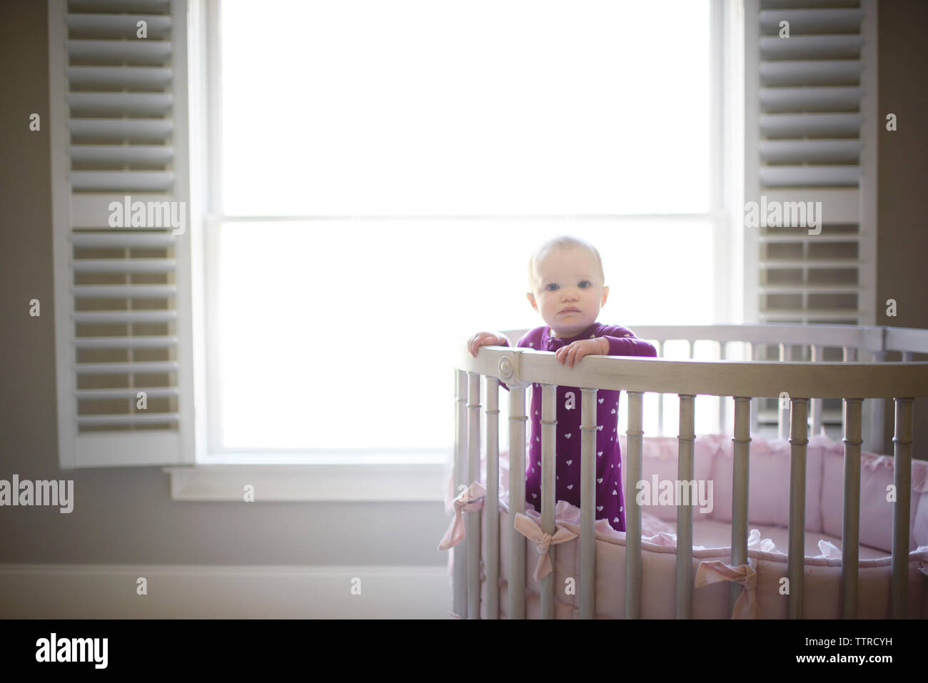 Portrait Of Cute Baby Girl Standing In Crib Against Window At Home