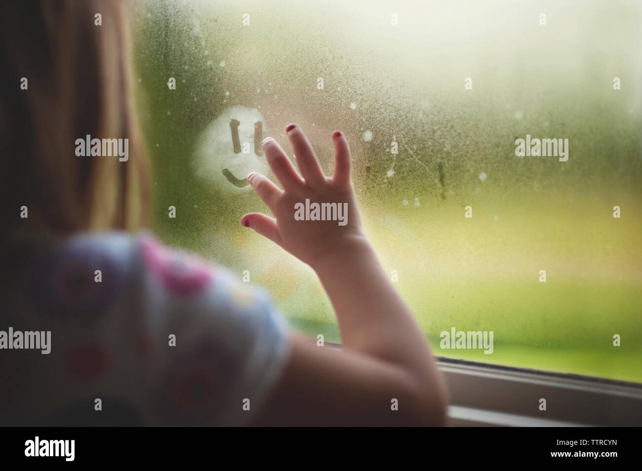 Cropped image of girl making anthropomorphic smiley face on wet window at home during rainy season Stock Photo