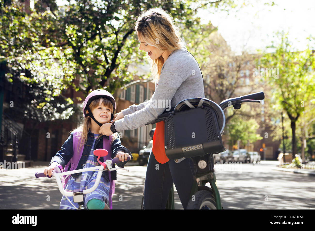 Woman assisting daughter in wearing cycling helmet on street Stock Photo