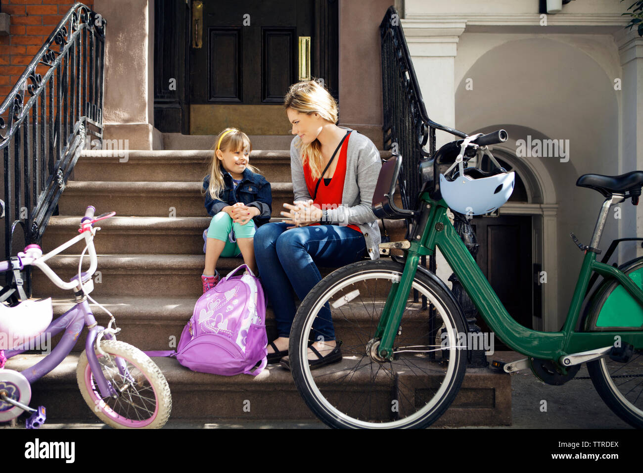Mother and daughter talking while sitting on steps with bicycles parked in foreground Stock Photo