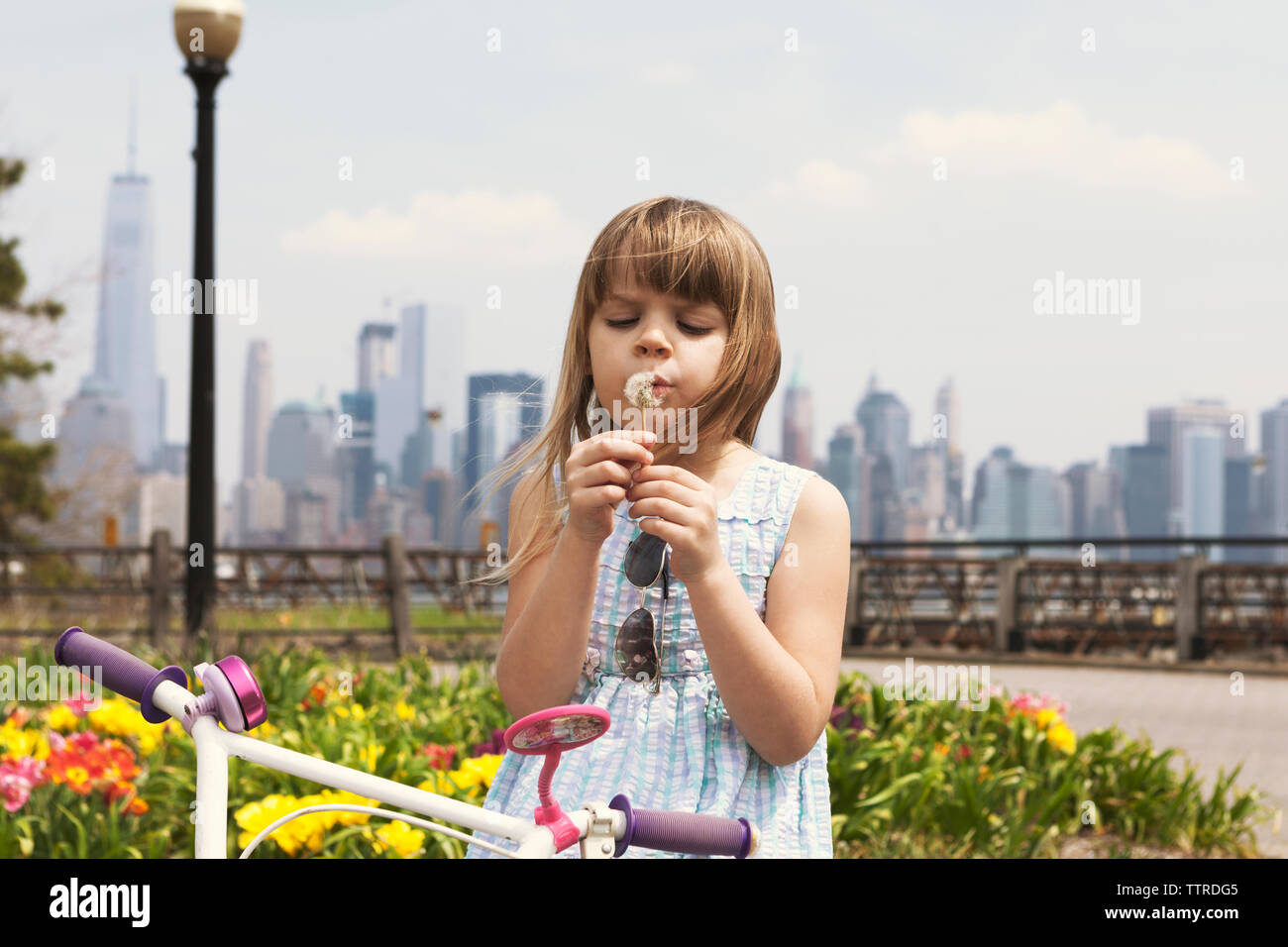 Cute girl blowing dandelion on promenade with city skyline in background Stock Photo