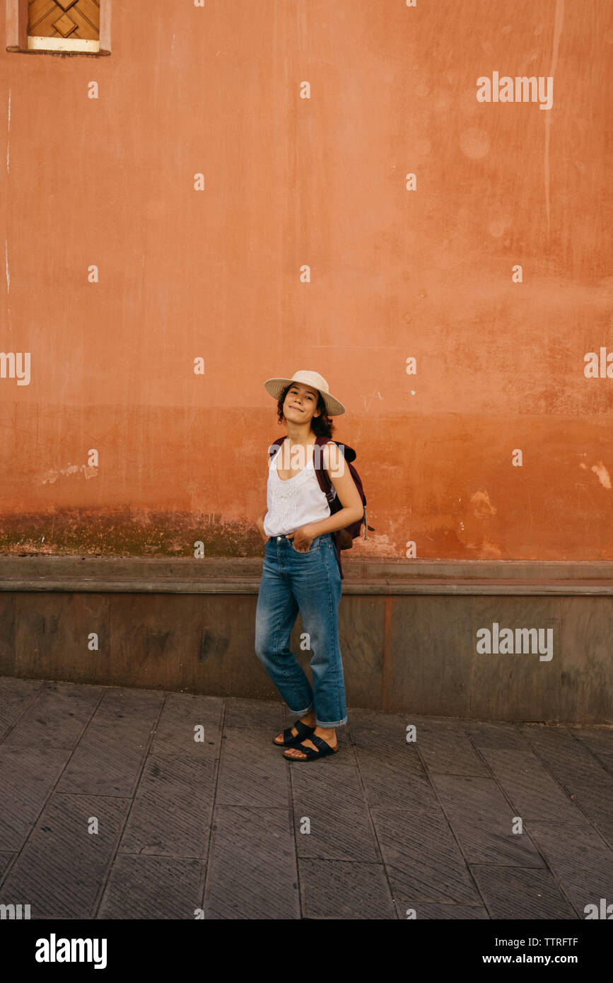 Full length portrait of confident female tourist with backpack and hands in pockets standing against wall in city Stock Photo