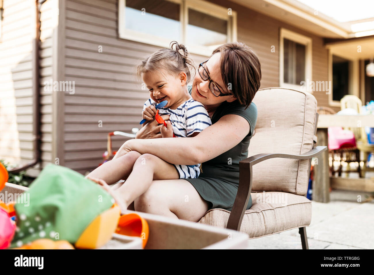 happy-daughter-with-toys-sitting-on-mother-s-lap-at-backyard-stock