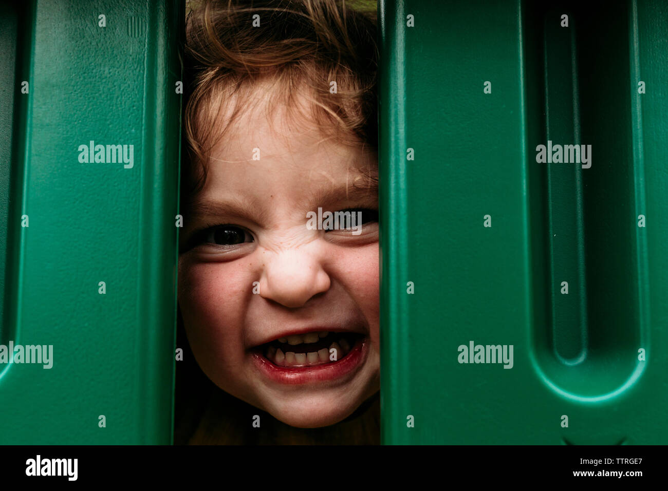 Portrait of cute playful girl making face while peeking through outdoor play equipment Stock Photo