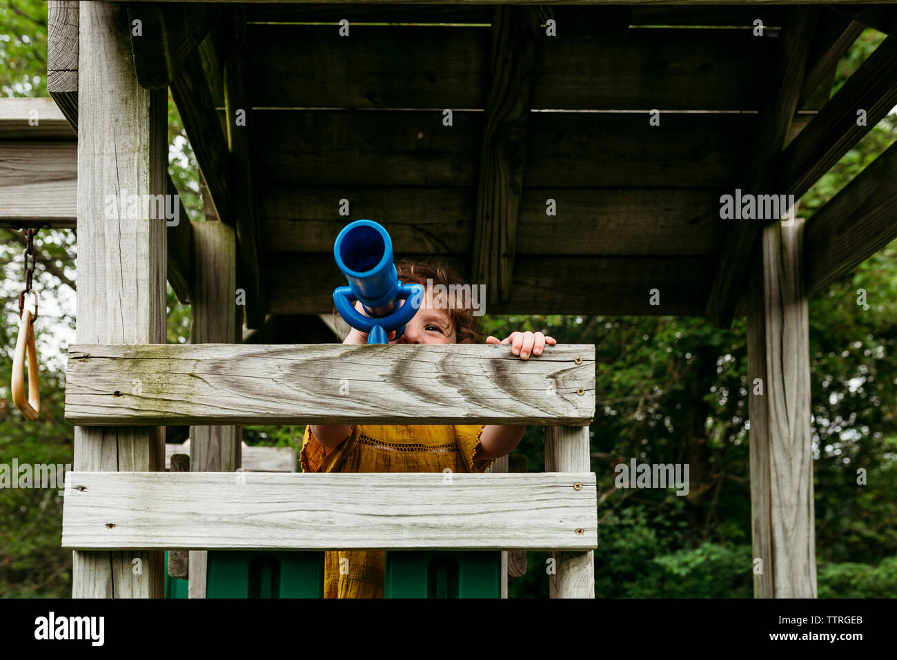 Girl looking through telescope while standing on wooden outdoor play equipment at playground Stock Photo