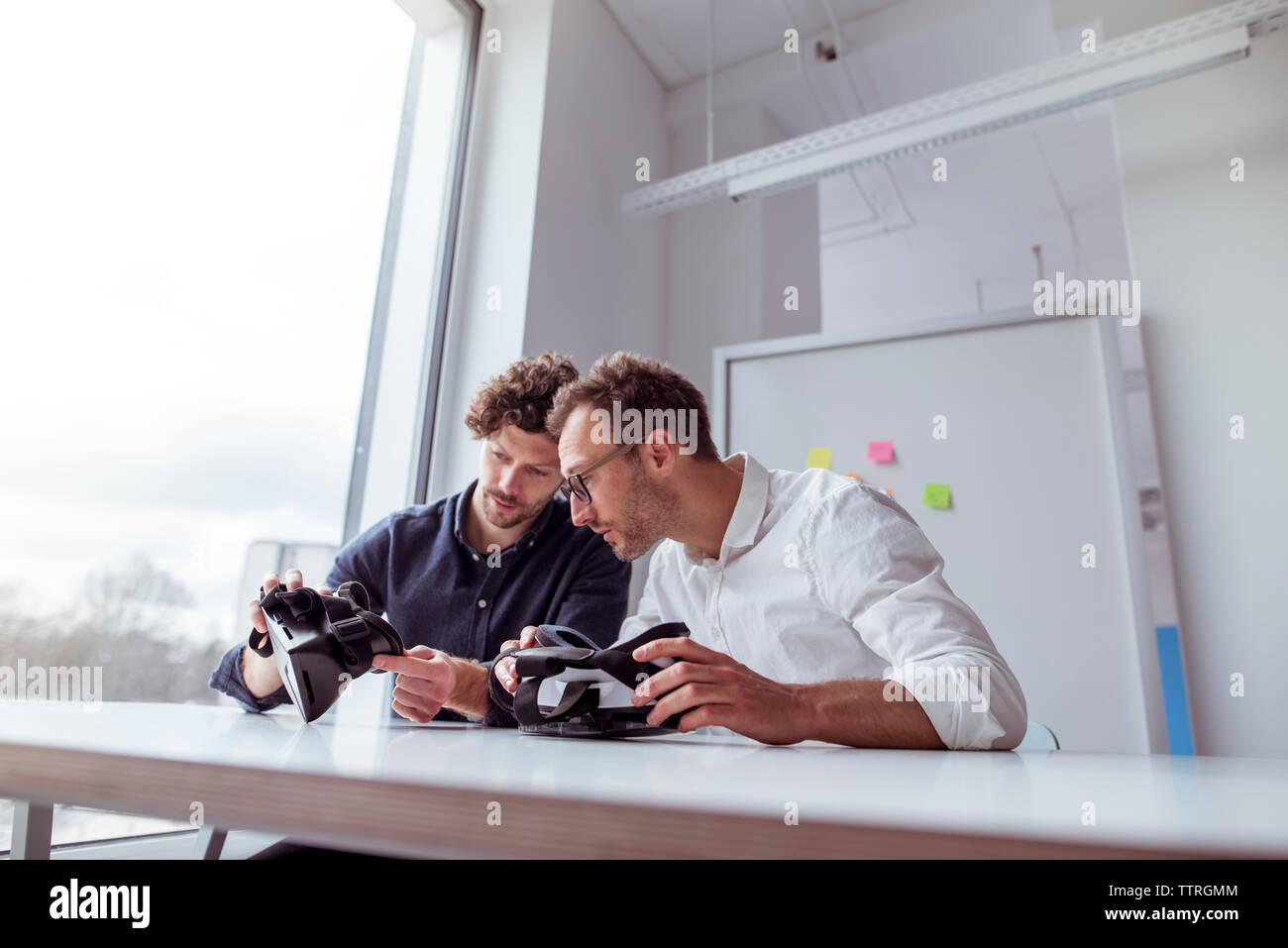 Technicians examining virtual reality simulators while sitting by window at table Stock Photo