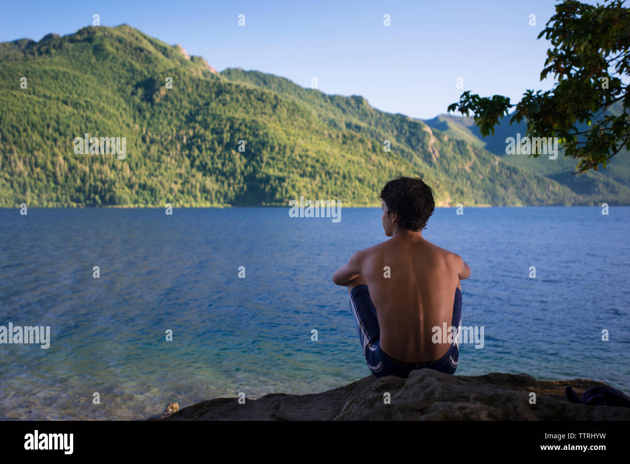 Rear view of teenage boy sitting on rock looking at lake and mountains ...