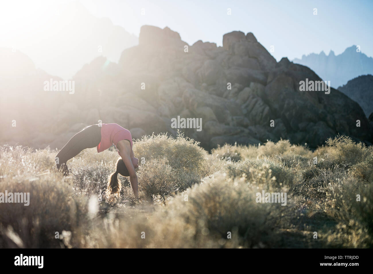 Woman bending over backwards while practicing yoga on field against mountains during sunny day Stock Photo