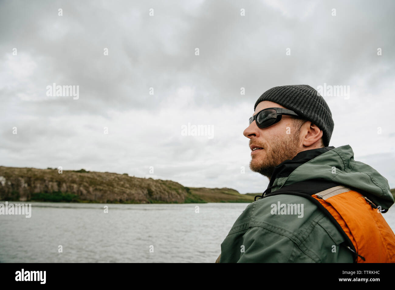 Side view of man wearing sunglasses and knit hat while standing by lake against cloudy sky at Iceland Stock Photo