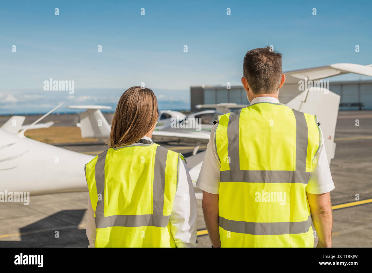 Rear view of engineers in reflective clothing standing against blue sky at airport runway Stock Photo