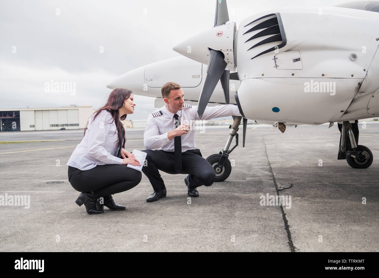 Engineer showing airplane parts to female trainee while crouching on airport runway against cloudy sky Stock Photo