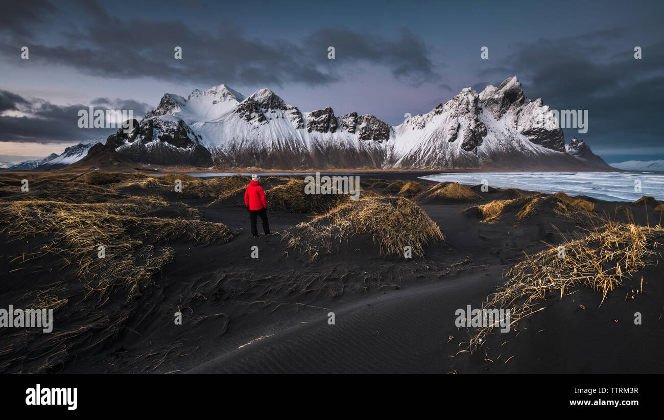Back view of person on dark ground of wild lands near river and high rock hills in snow in Vestrahorn, Stokksnes, Iceland Stock Photo