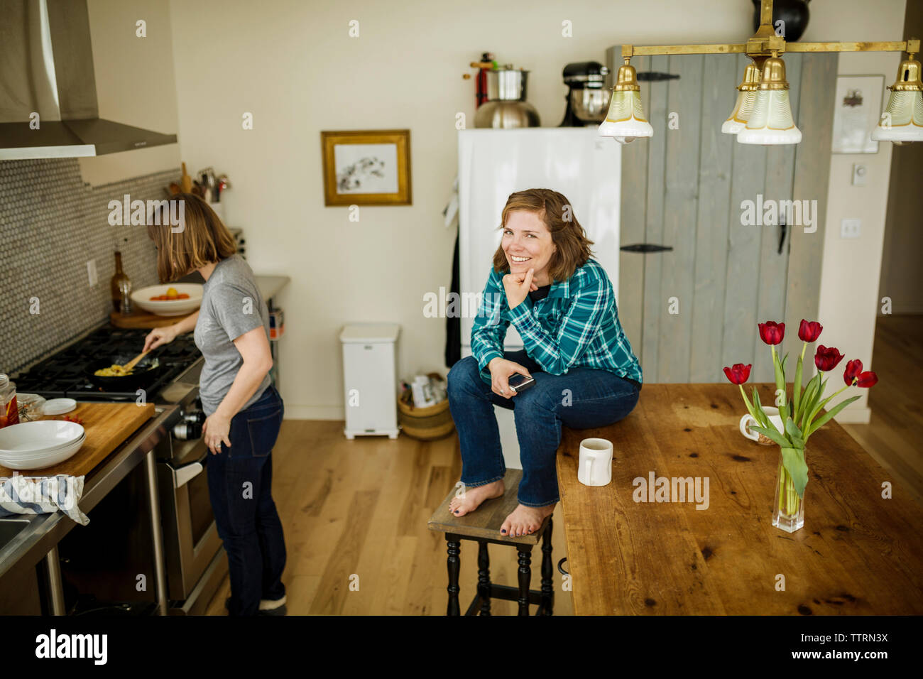 Woman sitting on table by sister preparing food at home Stock Photo