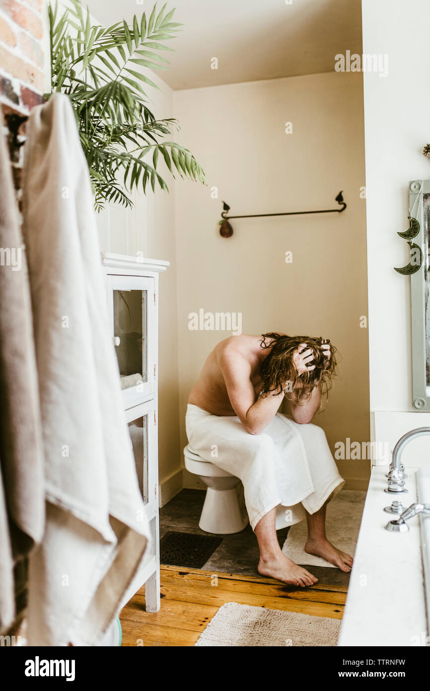 Depressed shirtless man with head in hands sitting on toilet bowl at
