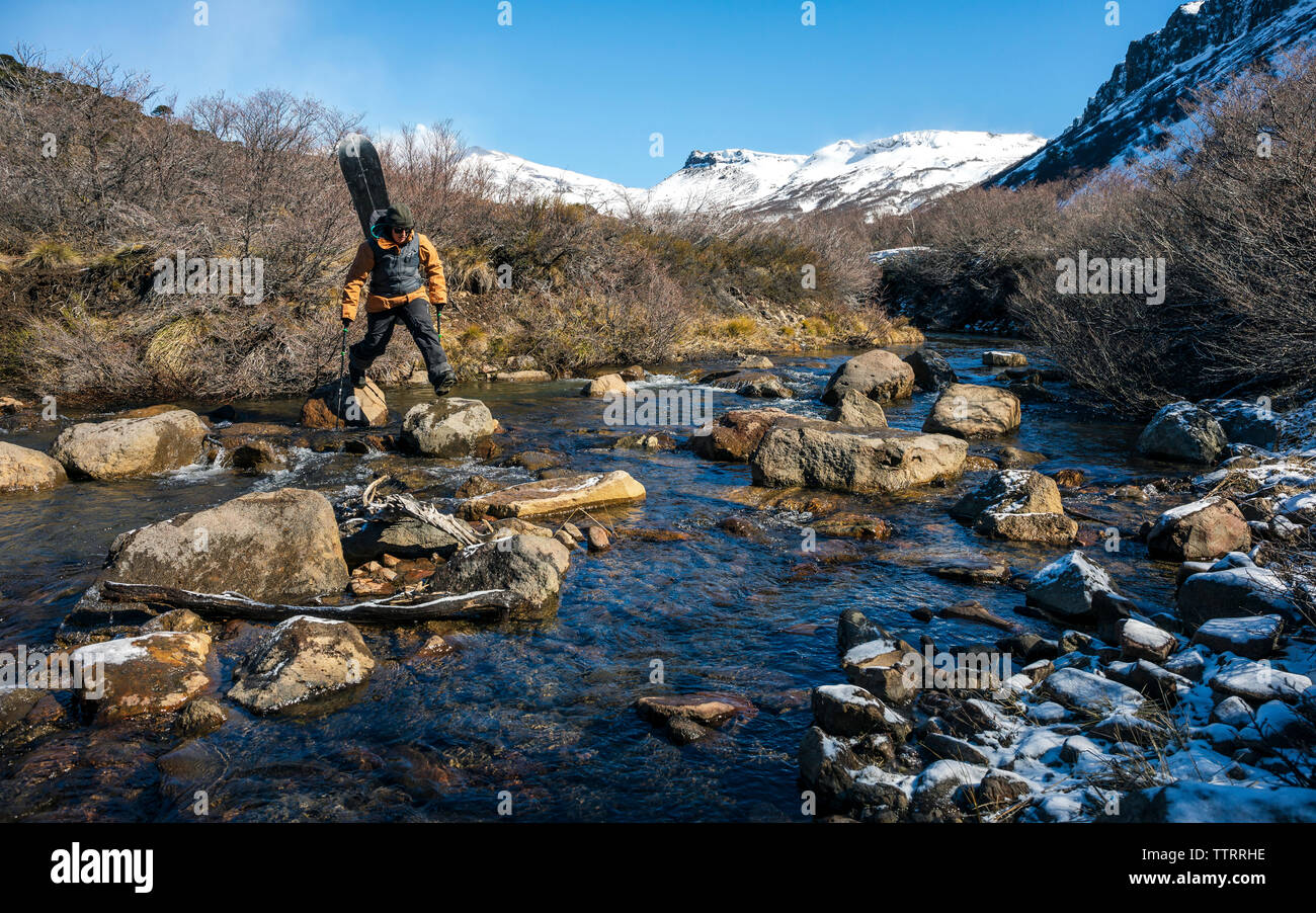 A women who is carrying a splitboard on her back hops across a creek on a search to find snow on a mission in Argentina. Stock Photo