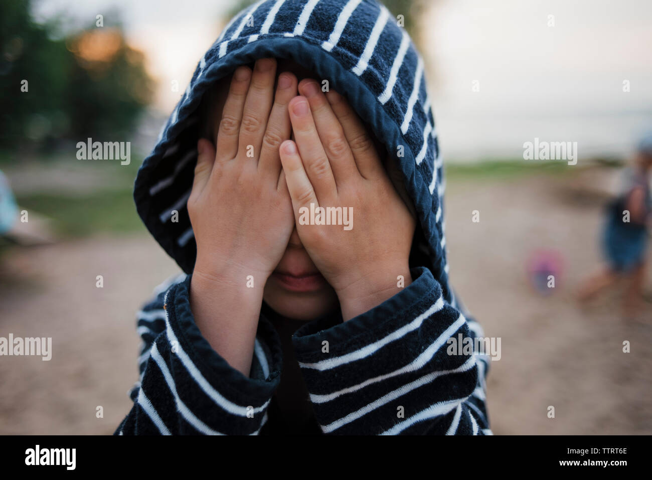 Boy in hooded shirt with hands covering face at beach Stock Photo