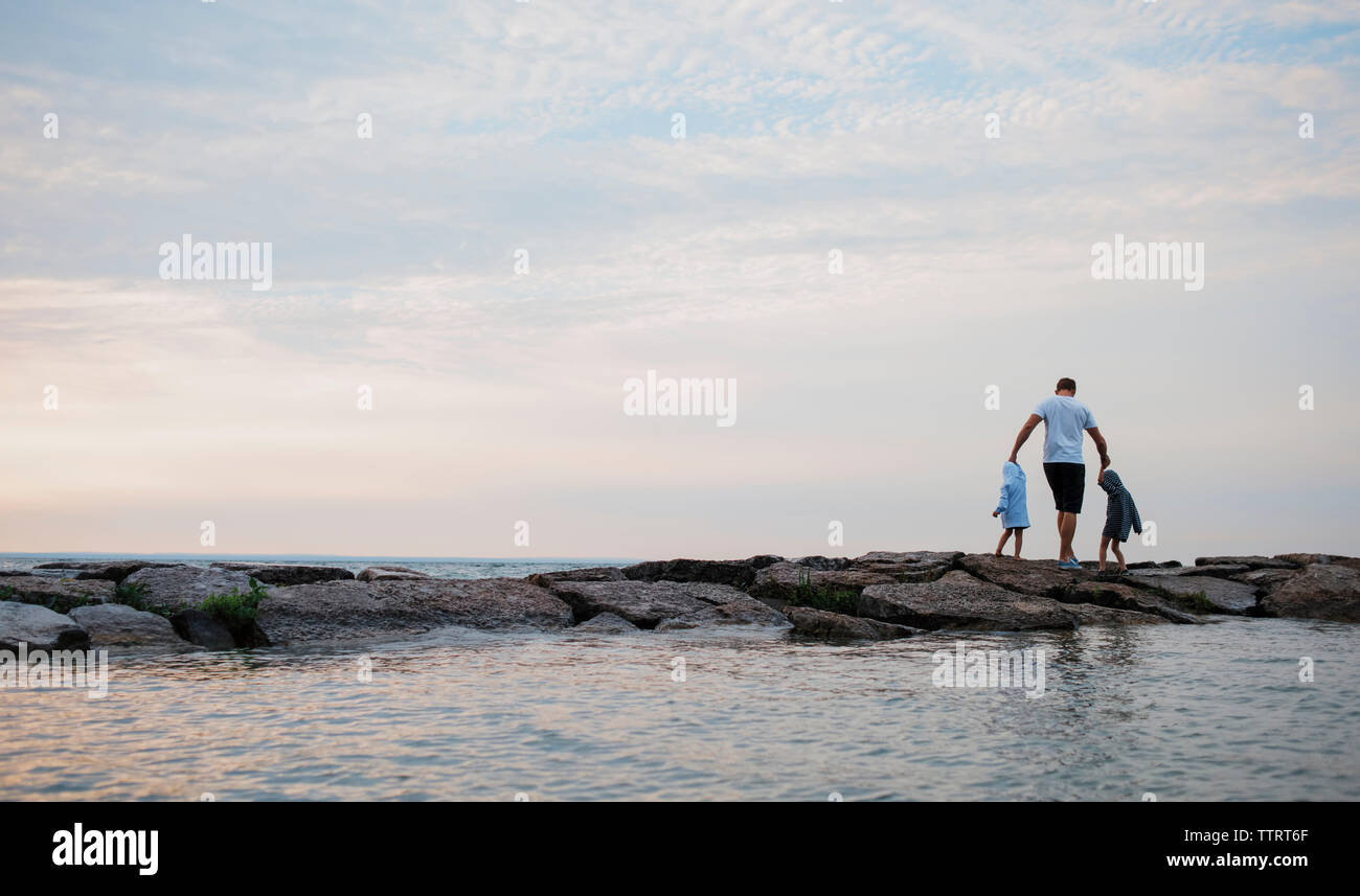 Rear view of father and children enjoying by sea against cloudy sky Stock Photo