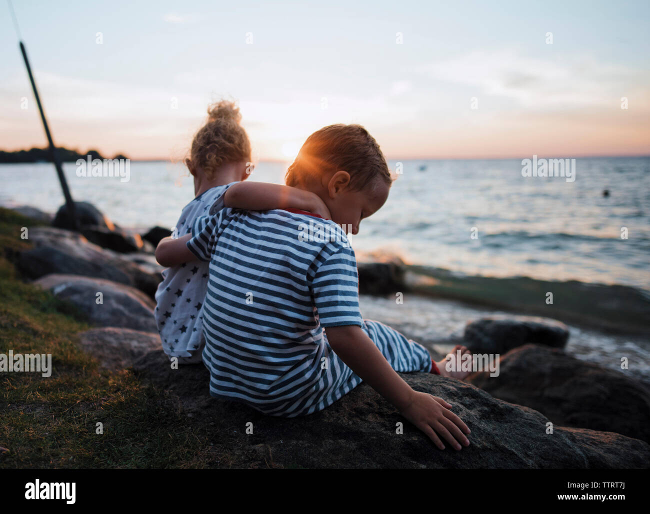 Siblings sitting on rocks by Lake Simcoe against sky during sunset Stock Photo