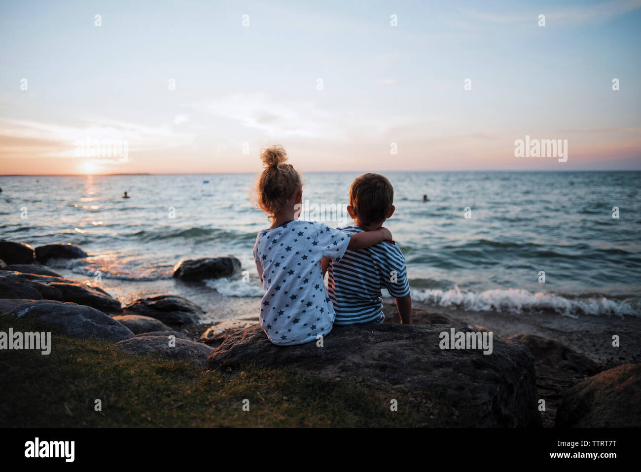 Rear view of siblings sitting on rocks by Lake Simcoe against sky during sunset Stock Photo