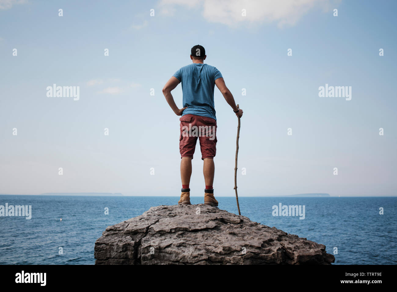 Rear view of hiker with hand on hip holding stick while standing on rock against sea and sky at Bruce Peninsula National Park Stock Photo