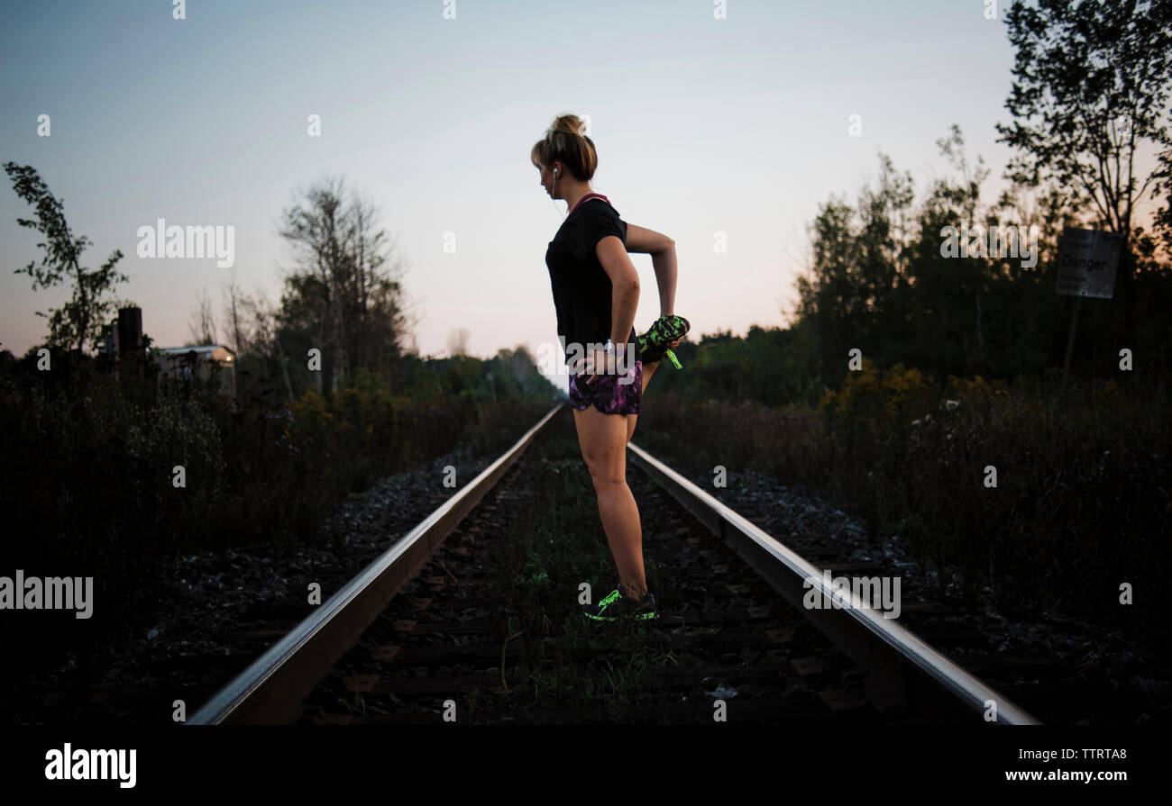 Side view of woman stretching leg while standing on railroad track against sky during dusk Stock Photo