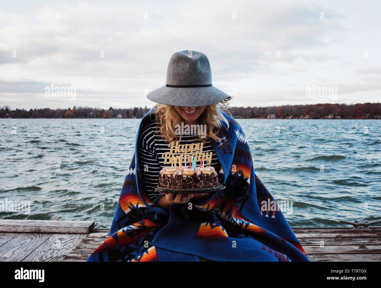Woman holding birthday cake while sitting on pier by lake against cloudy sky Stock Photo