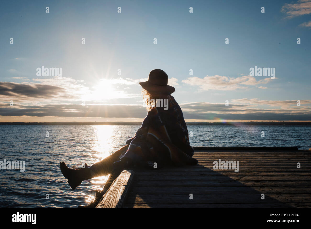 Side view of woman wrapped in blanket sitting on pier by Lake Simcoe during sunset Stock Photo